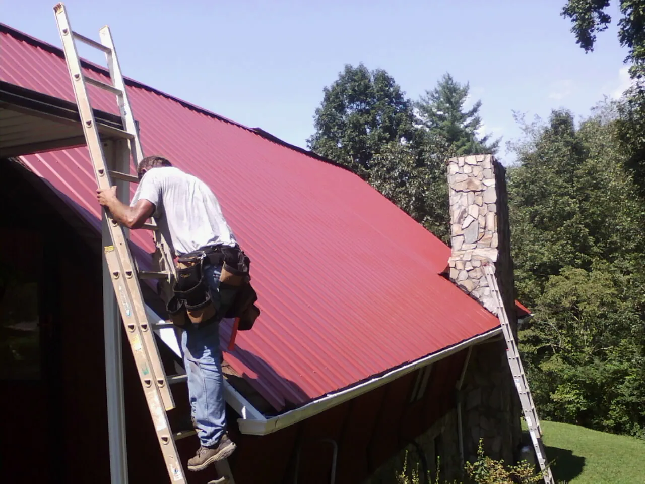 Roofer on a ladder beside a red metal roof and stone chimney — Soffit & Fascia Repair by Roofing Brothers in La Crosse